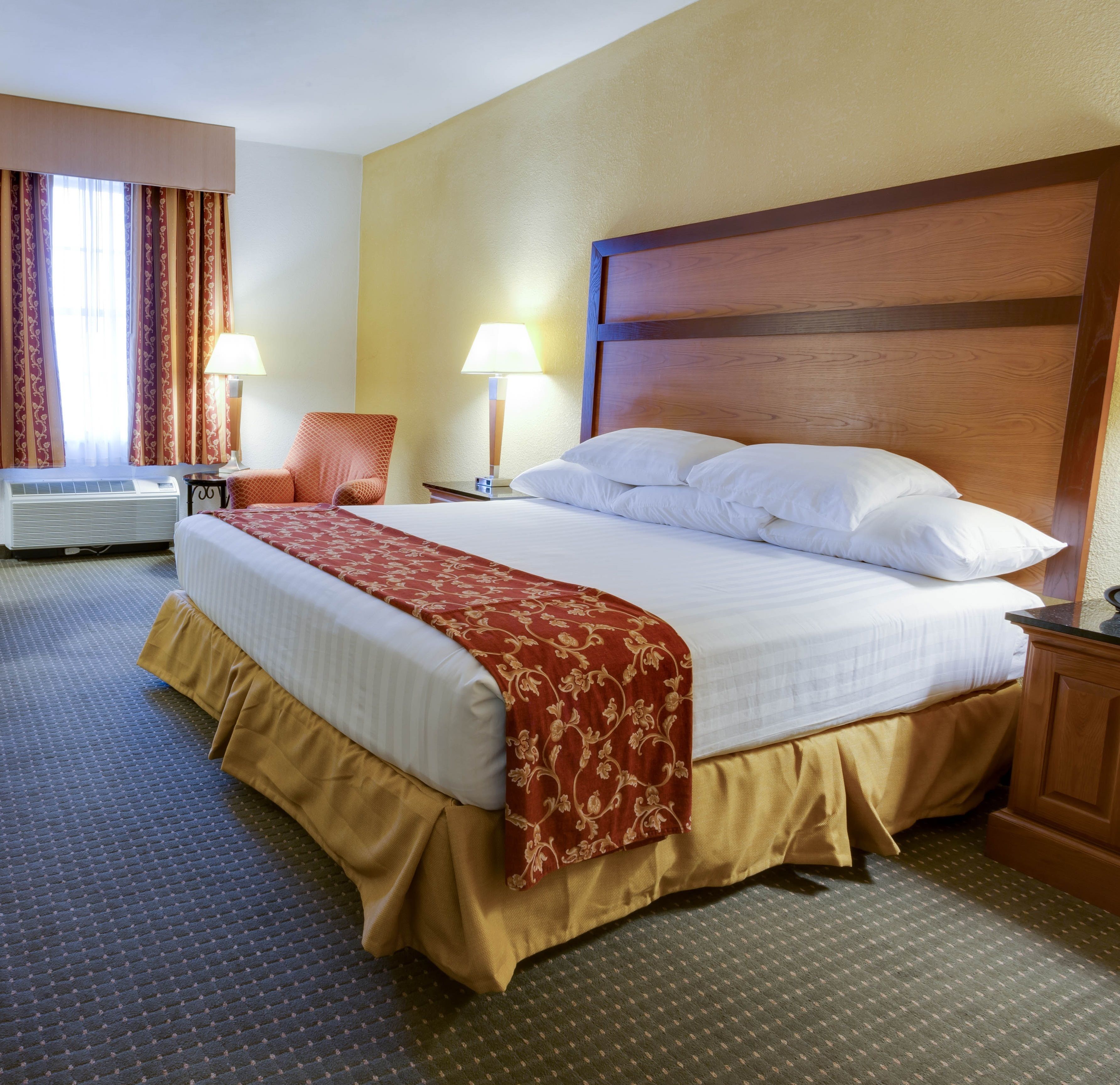 A hotel room with a large bed, two bedside lamps, a chair, and an air conditioning unit.