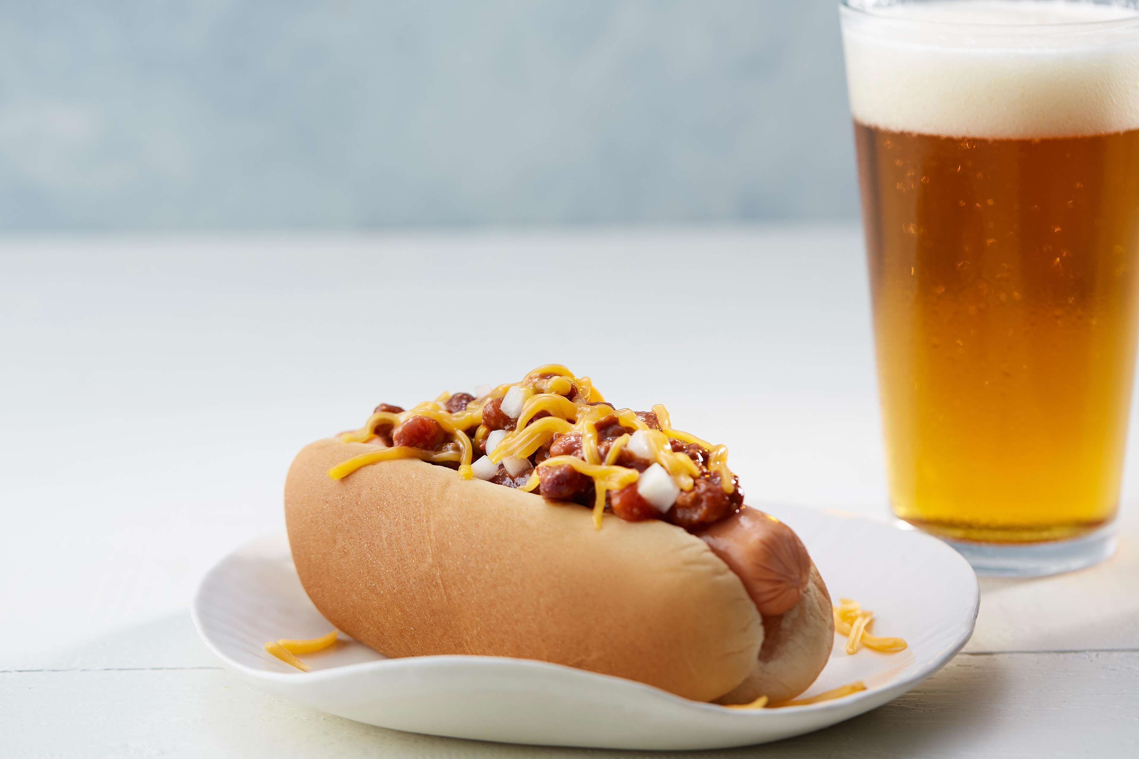 A hot dog with chili and cheese on a bun, served on a white plate, with a glass of beer in the background.
