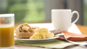 Desayuno con waffles, huevos revueltos, tostadas, un vaso de jugo de naranja y una taza blanca sobre una mesa con un mantel individual verde.