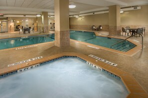 Een binnenzwembad met een jacuzzi, betegelde vloer en een zitgedeelte met tafels en stoelen.