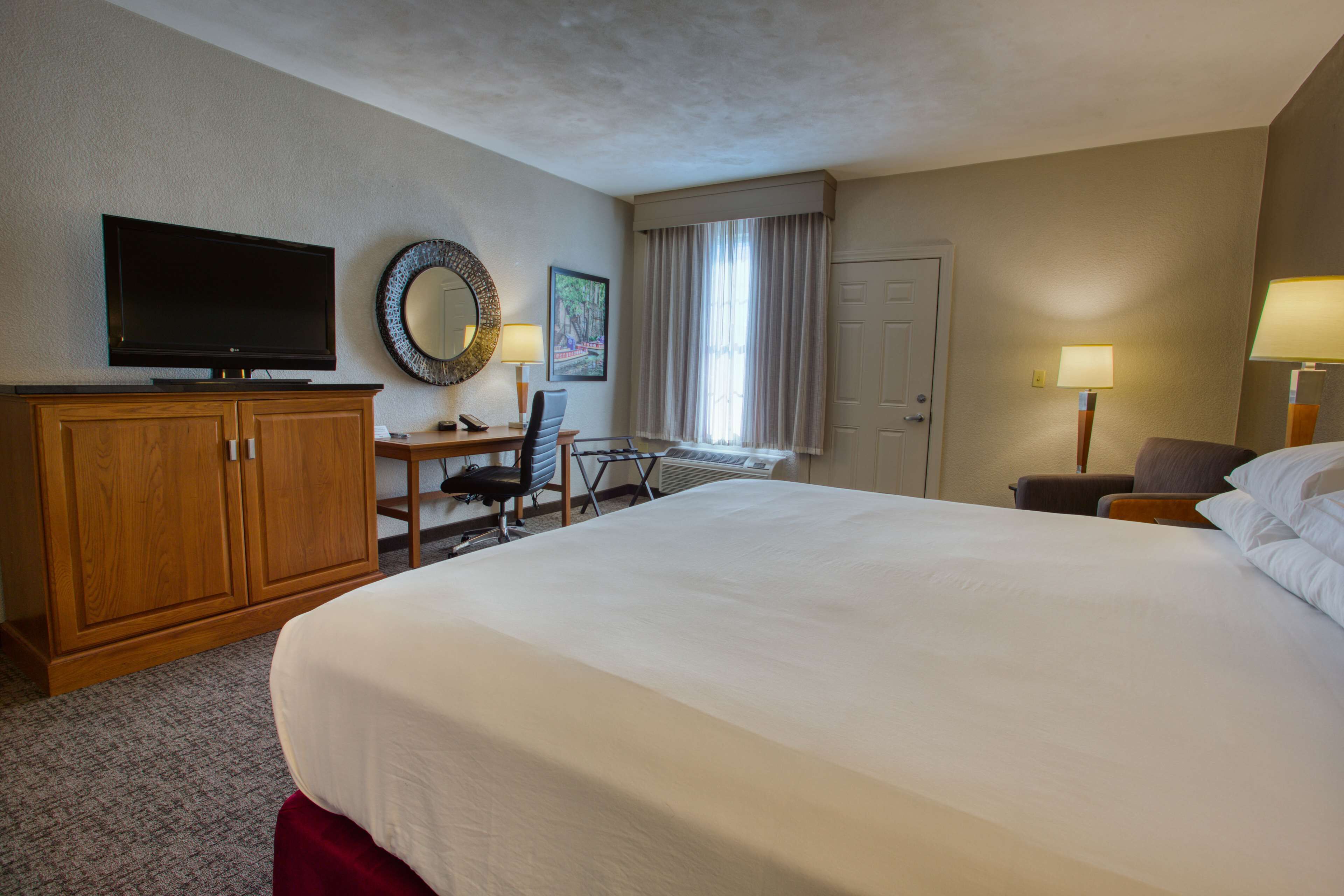 A hotel room with a large bed, a desk with a chair, a television, a mirror, and two lamps.