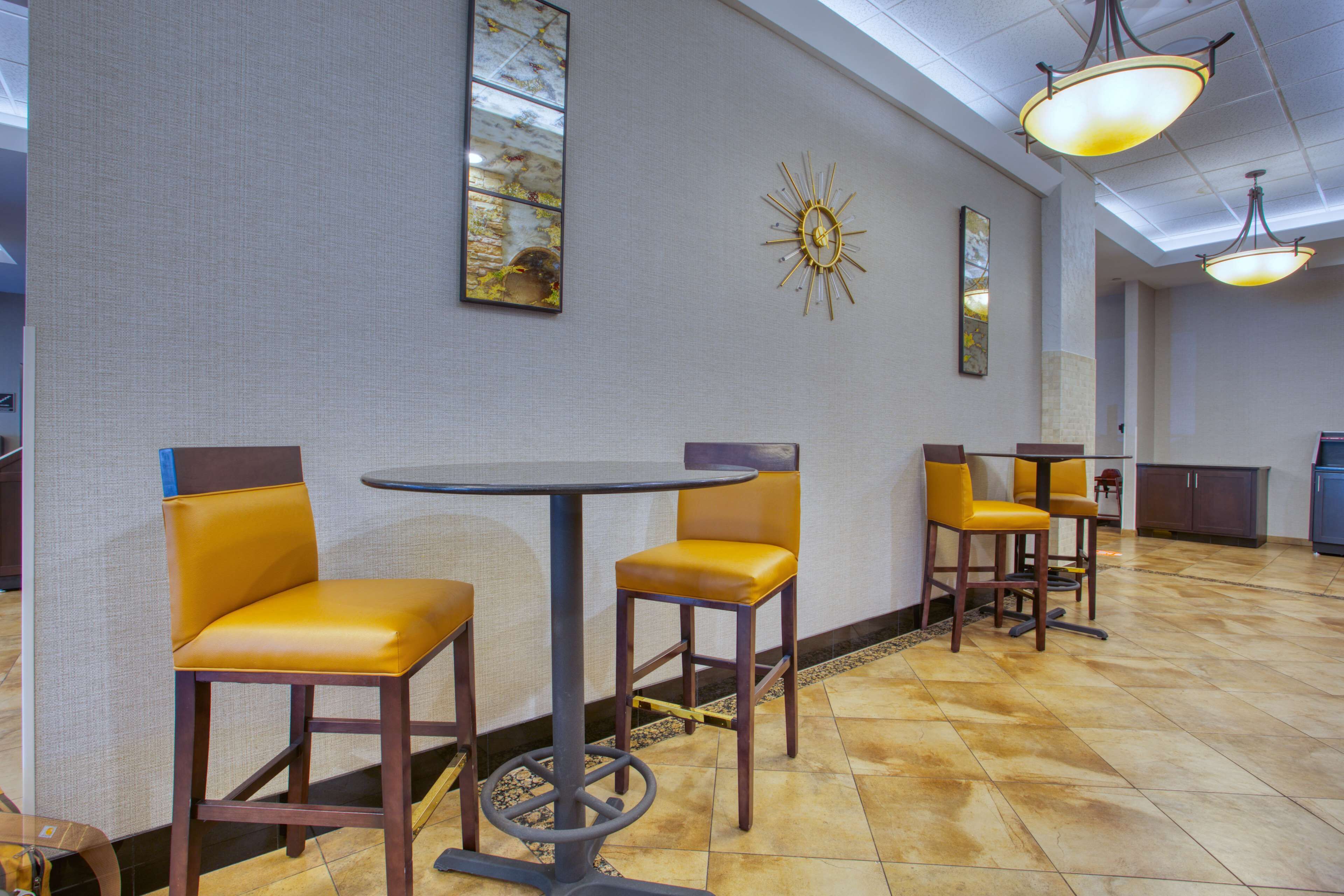 A hotel lobby with a round table and four yellow chairs, a decorative wall clock, and a ceiling light fixture.