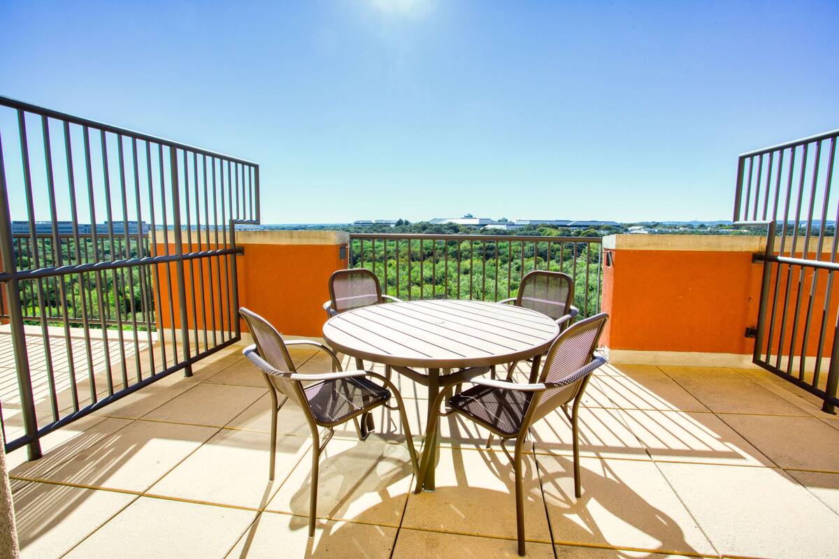 Un balcon doté d’une table ronde et de quatre chaises, entouré de murs orange et de rampes en métal, offrant une vue sur un paysage verdoyant.