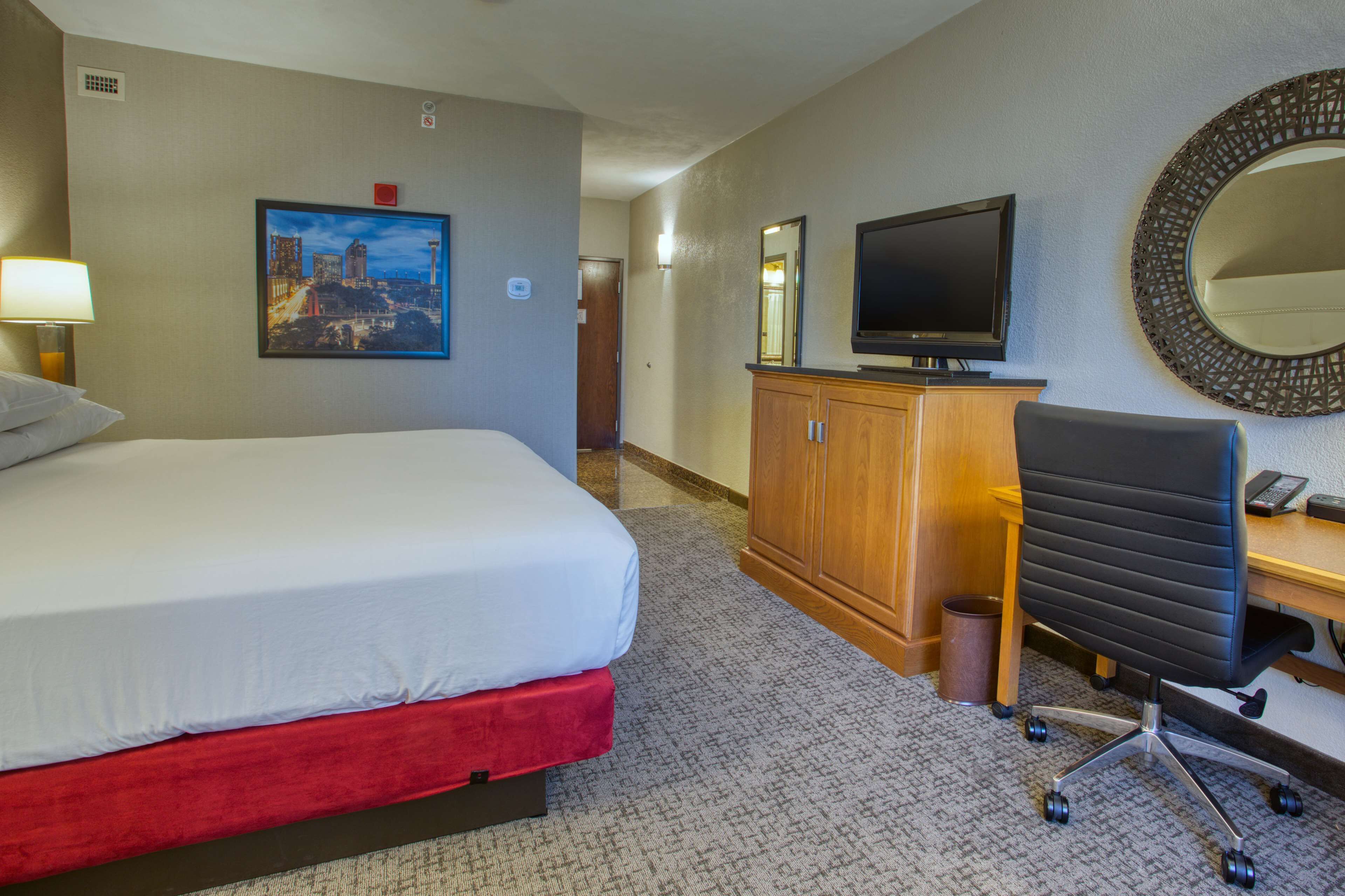 Een hotelkamer met een bed, een bureau met stoel, een televisie, een spiegel en een schilderij aan de muur.