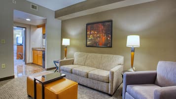 Habitación de hotel con sofá, sillón y reposapiés, un cuadro en la pared y una lámpara a cada lado del sofá.
