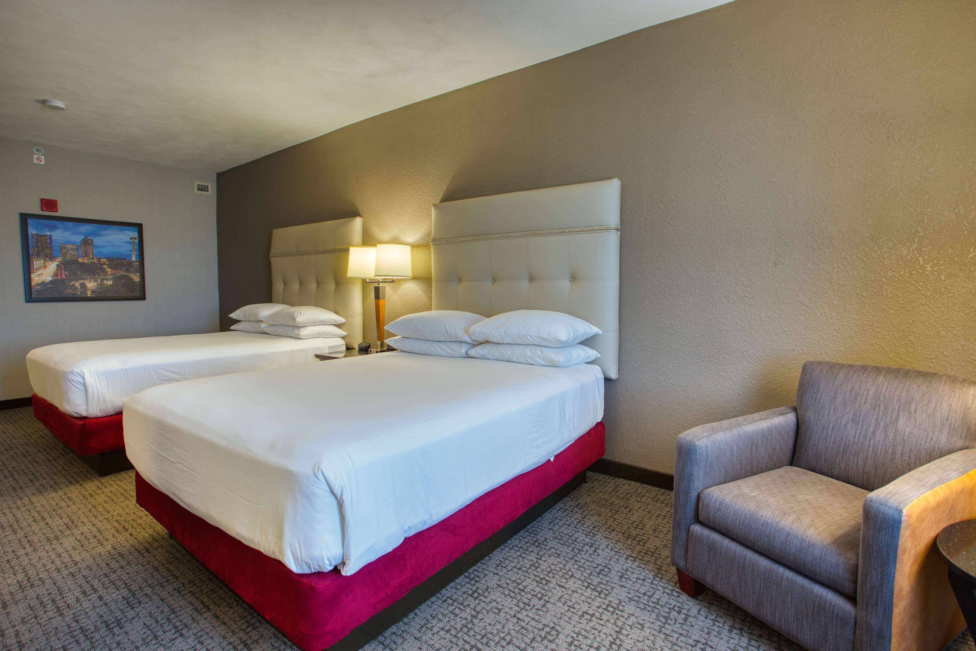 Habitación de hotel con dos camas, un detalle rojo en la cama inferior, un sillón gris y una lámpara en la mesita de noche.