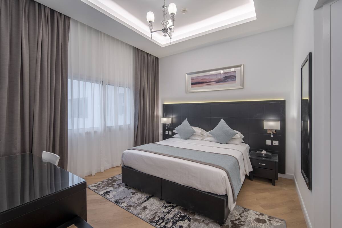 A modern hotel room with a large bed, bedside tables, a desk, and a chandelier.