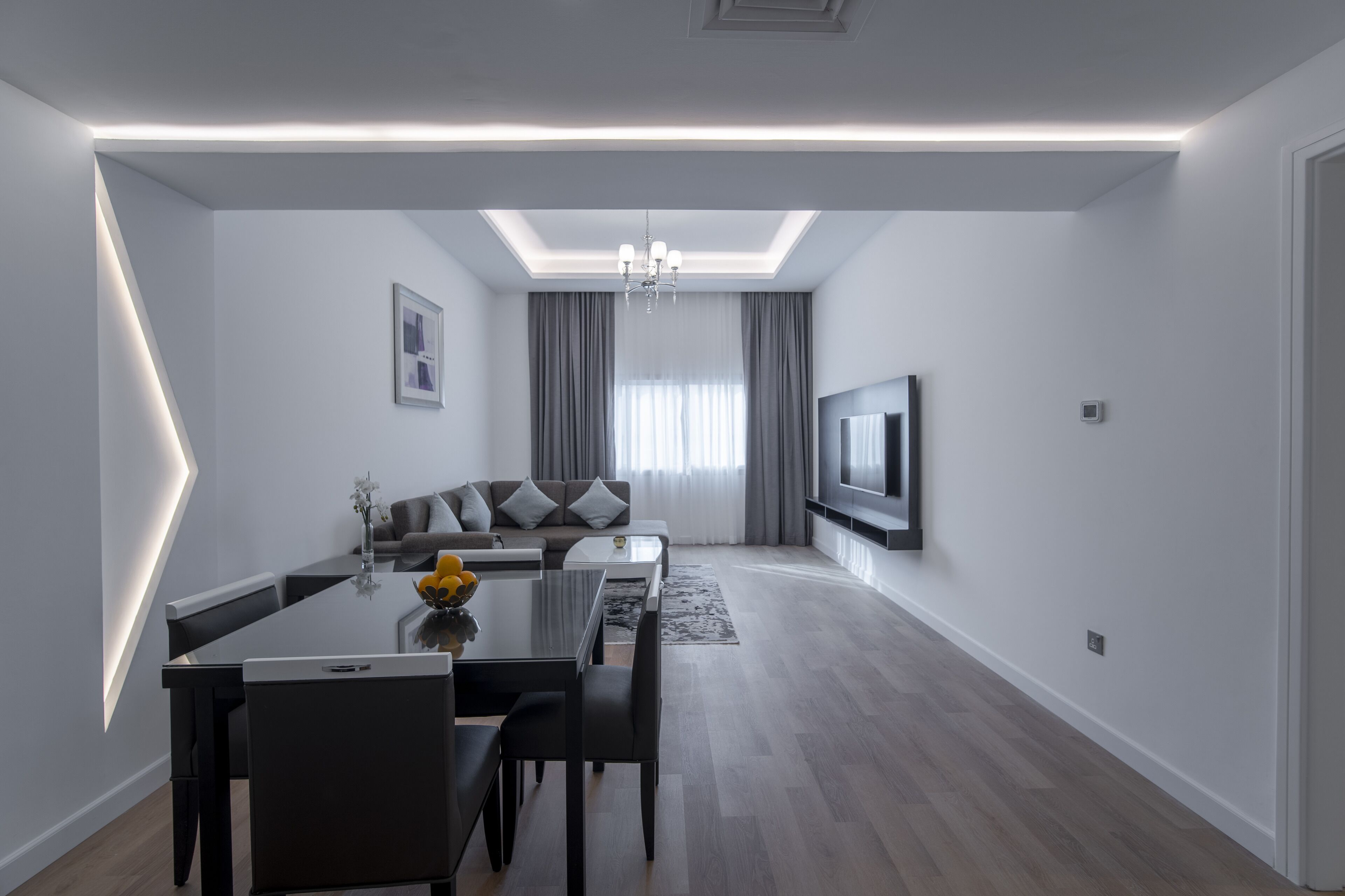 A modern living room with a dining area, a sofa set, a flat-screen TV, and a chandelier.