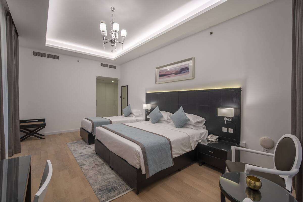 A modern hotel room with two beds, a desk, a chair, a small table, and a ceiling light fixture.