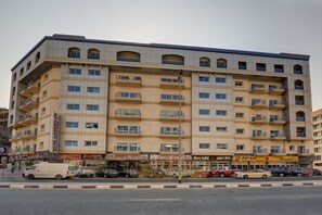 A multi-story building with balconies, a variety of shops on the ground floor, and several parked cars along the street. - Rose Garden Hotel Apartments Barsha (Dubai)