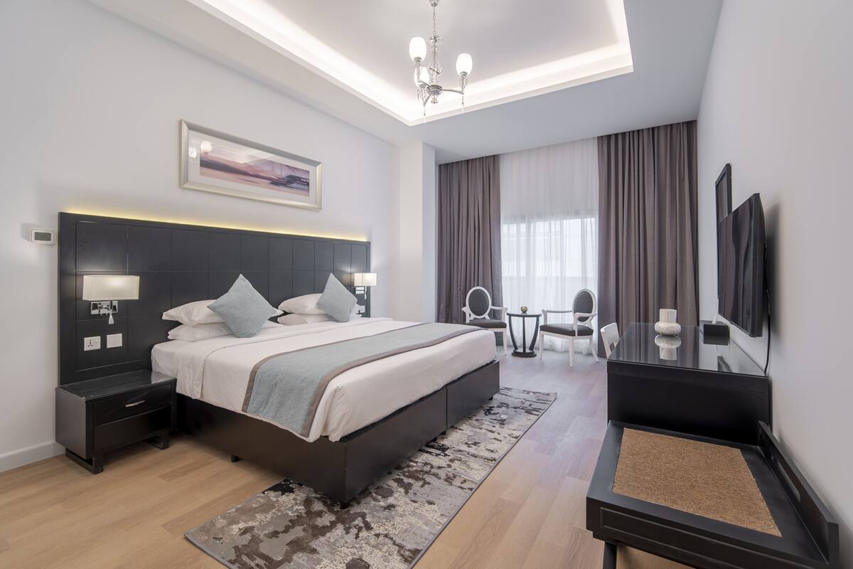 A modern hotel room with a large bed, bedside tables, a desk, a chair, a TV, and a large window with curtains.