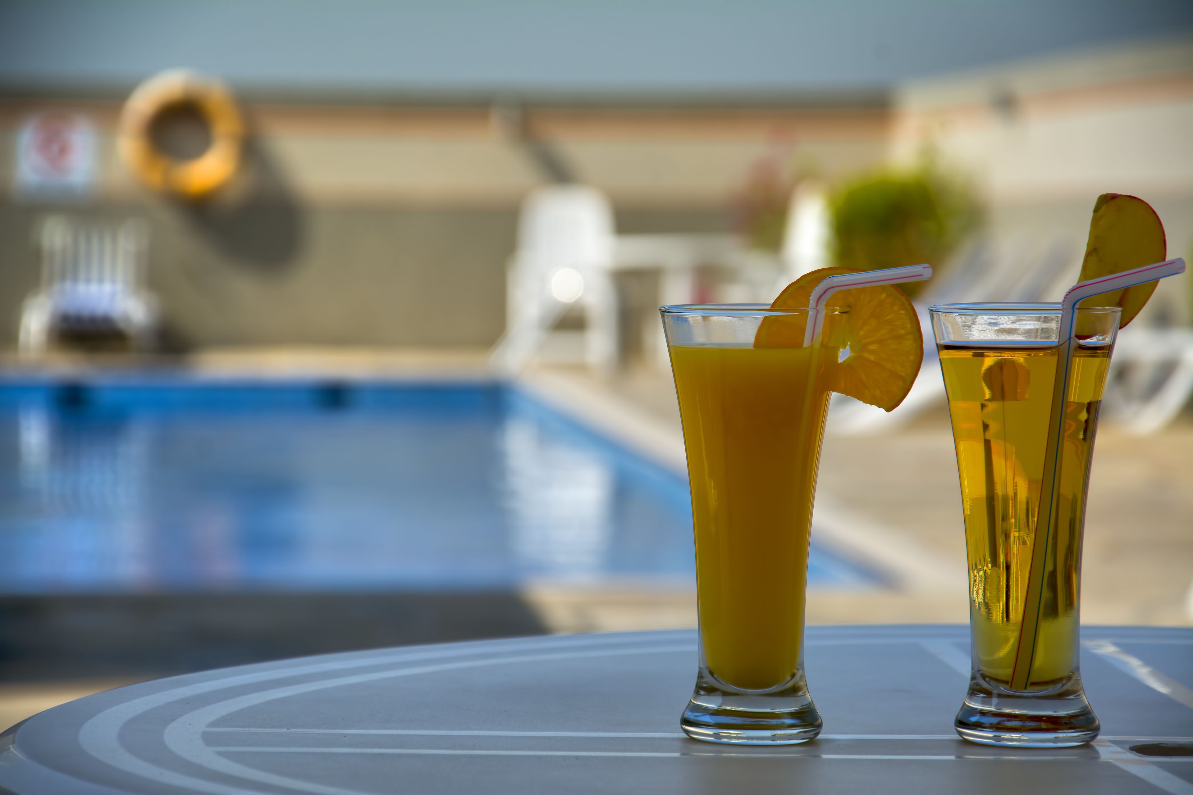 Two glasses of orange juice with straws and a slice of orange on a table by a pool.