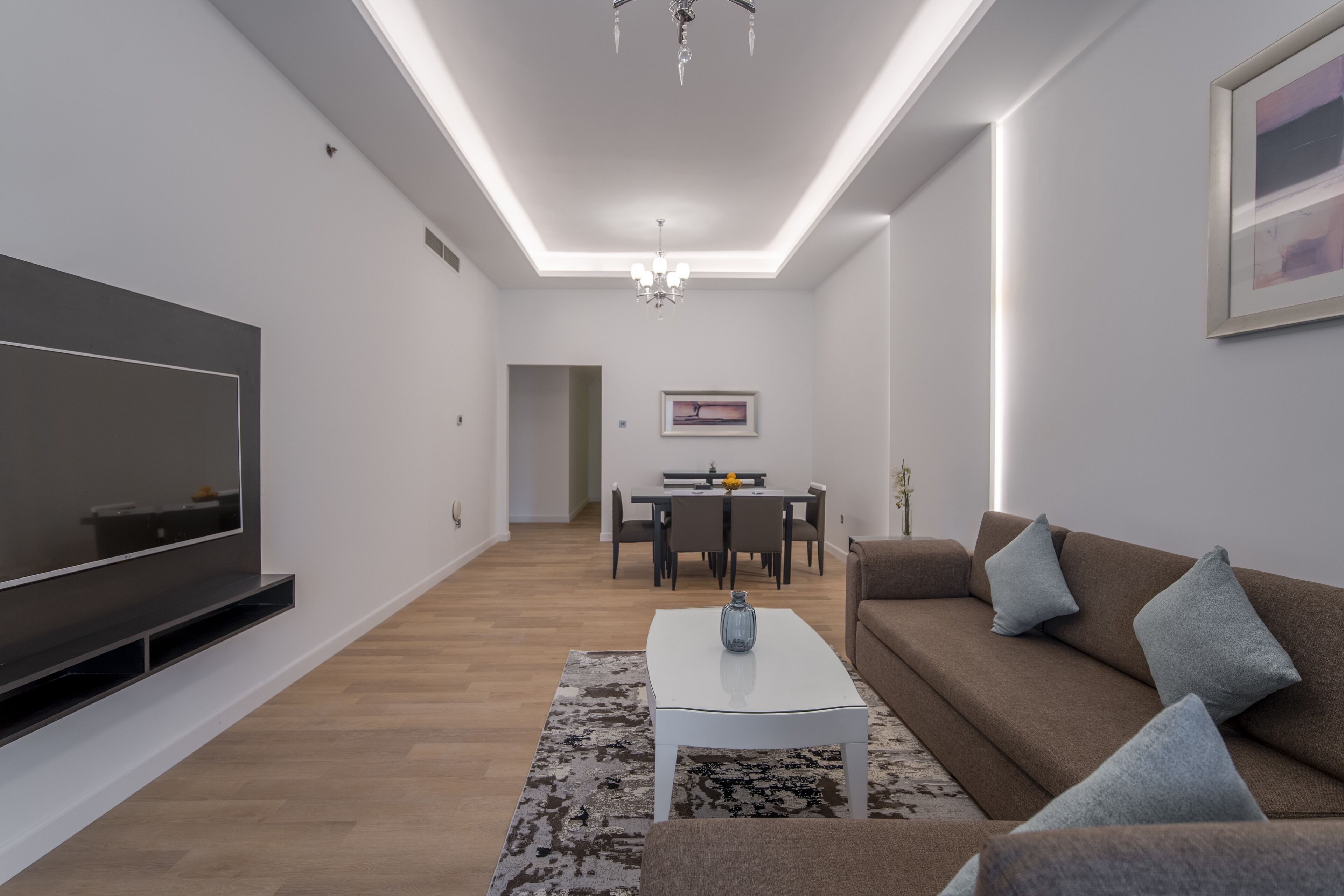 A modern living room with a flat-screen TV, a white coffee table, a brown sofa, and a dining area in the background.