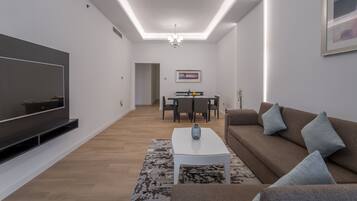A modern living room with a flat-screen TV, a white coffee table, a brown sofa, and a dining area in the background.