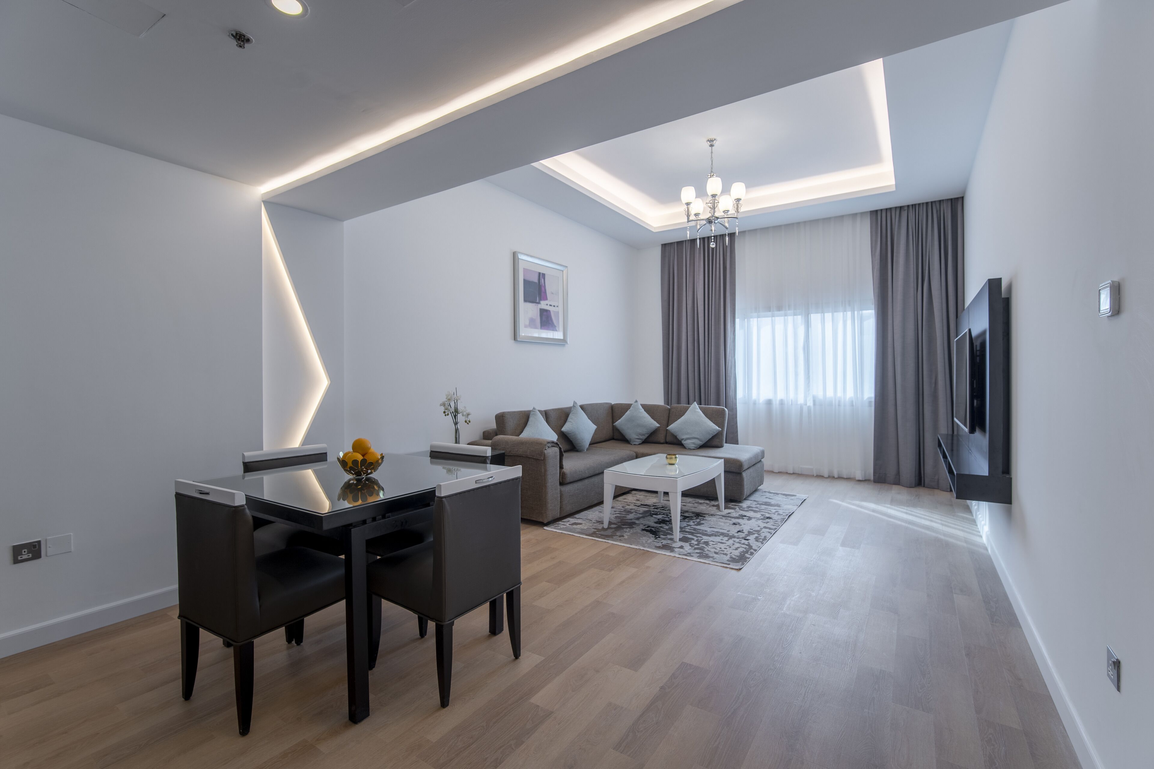 A modern living room with a dining area, a sofa set, a flat-screen TV, and a chandelier.