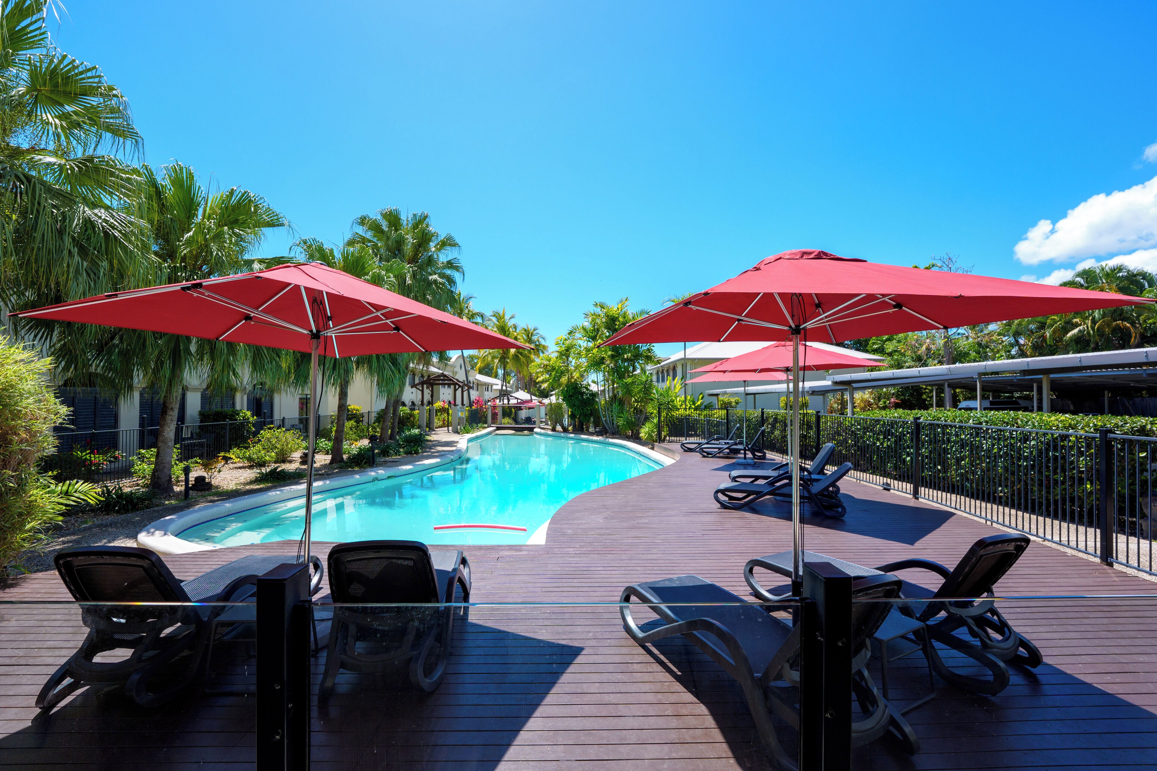 Un espace aménagé au bord de la piscine, avec des chaises longues et des parasols rouges, entouré de palmiers et surplombé d’un ciel bleu azur.