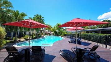 Un espace aménagé au bord de la piscine, avec des chaises longues et des parasols rouges, entouré de palmiers et surplombé d’un ciel bleu azur.