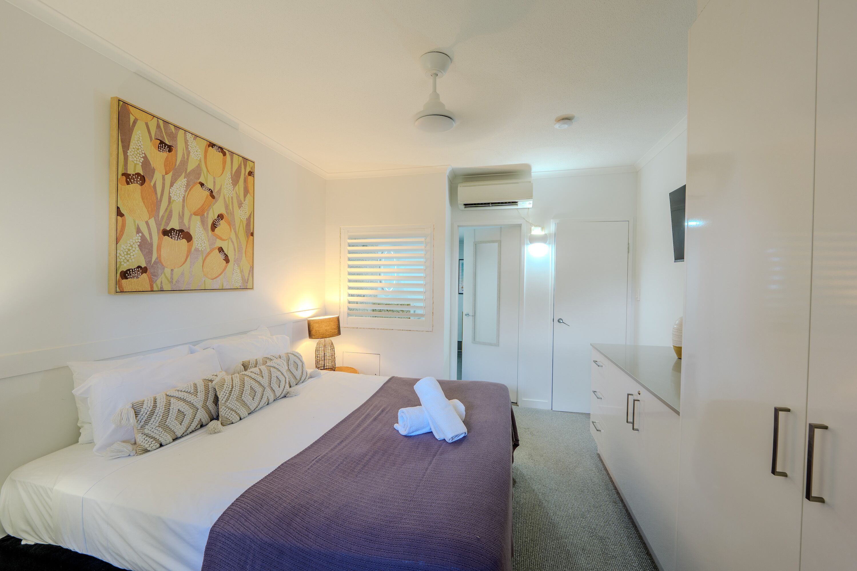 A hotel room with a bed, a large painting, a ceiling fan, an air conditioning unit, and a wardrobe.