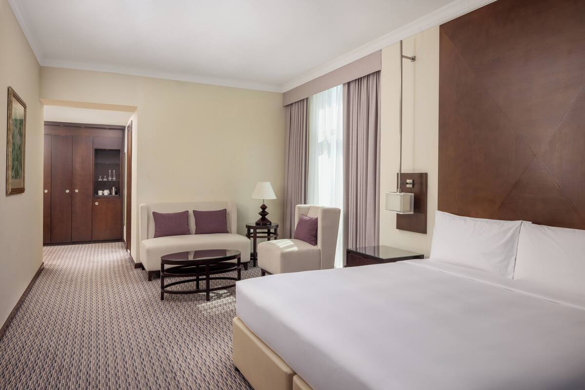A hotel room with a bed, two armchairs, a small table, a lamp, a nightstand, and a wardrobe.