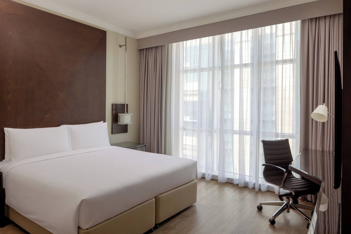 A hotel room with a large bed, a desk with a chair, a TV, and a window with curtains.