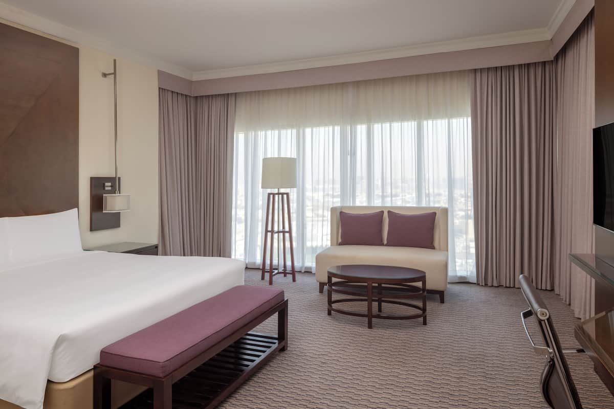 A hotel room with a bed, a sofa, a small table, a bench, a floor lamp, and a TV.