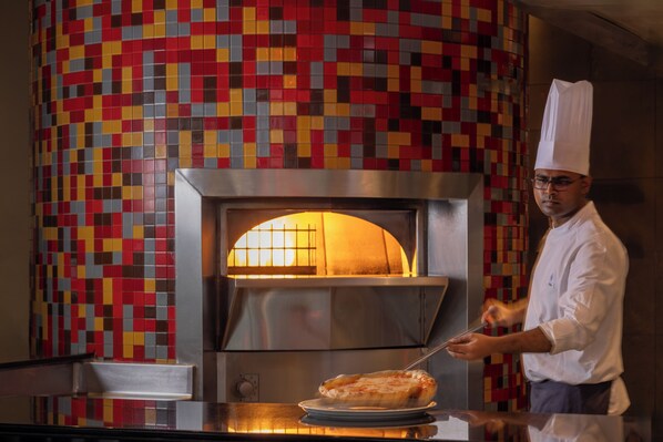 Un chef, vêtu d’un uniforme blanc et coiffé d’un haut-de-forme, prépare des plats devant un four en briques, le mur derrière étant décoré de carreaux colorés.