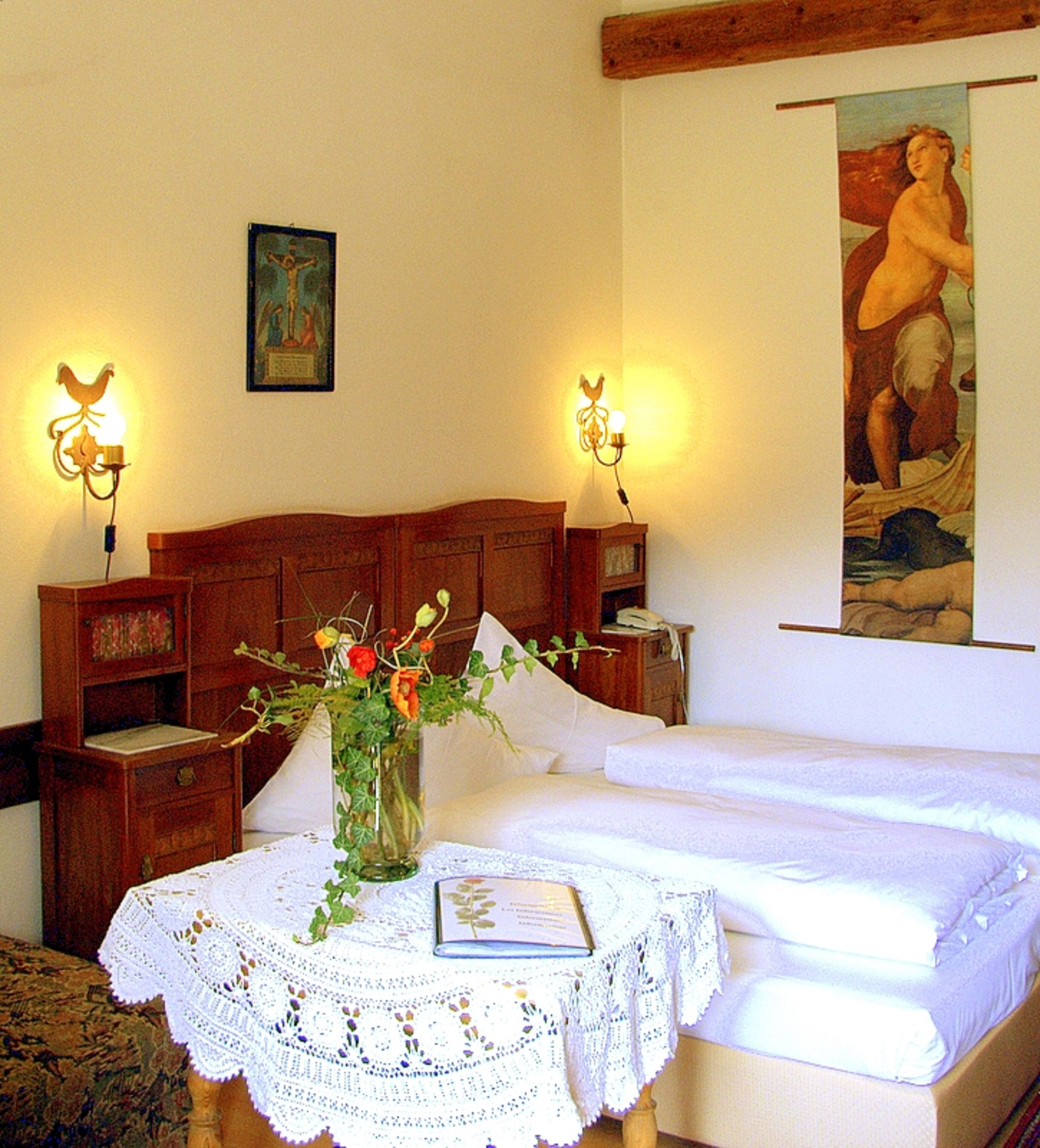 A bedroom with a wooden headboard, a white bedspread, a lace tablecloth, a vase with flowers, and two wall-mounted lamps.