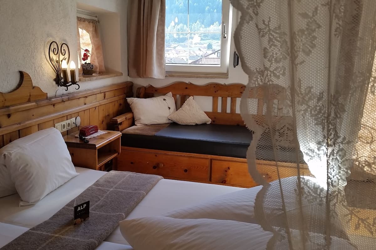 Habitación de hotel con cabecera de madera, cama con ropa blanca, mesita de noche con lámpara y ventana con vistas a un pueblo.