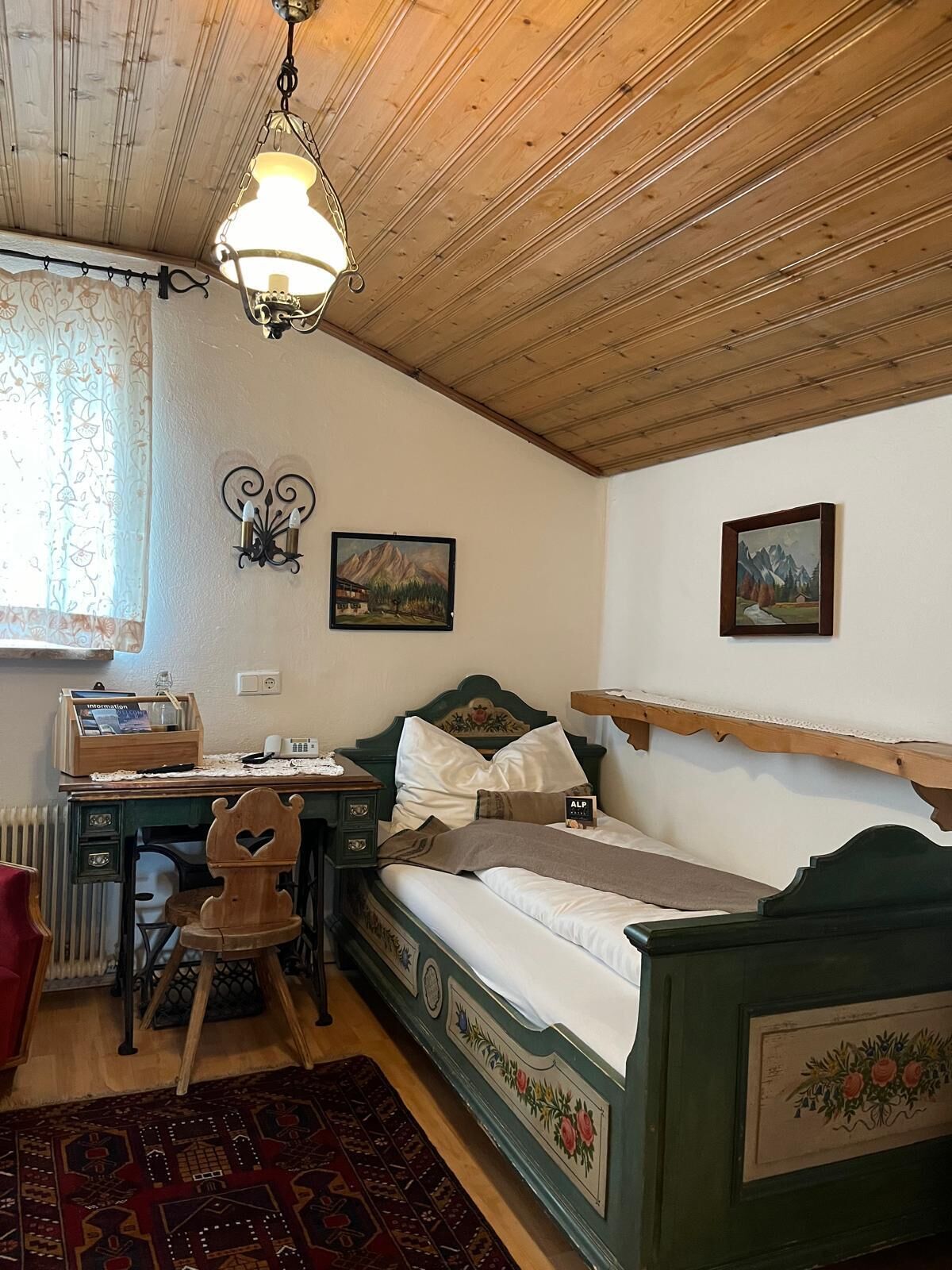 Ein Zimmer mit Holzboden, einem Bett mit grüner Kopfteil, einem Schreibtisch mit Stuhl, einem Heizkörper und einem Fenster mit Vorhängen.