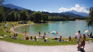 Un parco sul lago con persone che si prendono il sole e nuotano, circondato da alberi e montagne.