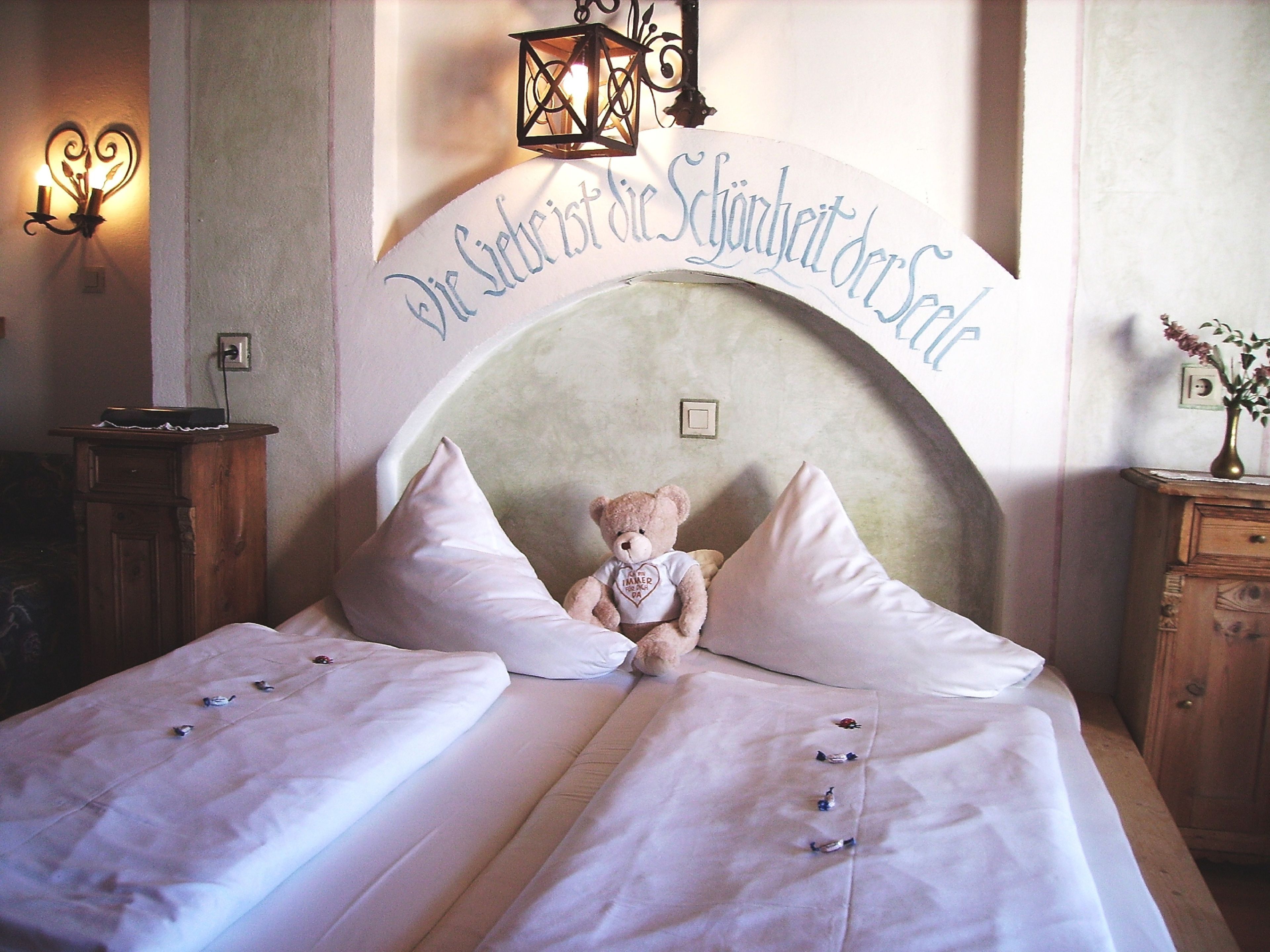 A neatly made bed with white linens and pillows, a teddy bear on the bed, and a sign above the bed that reads "Die Schönheit der Schweiz".