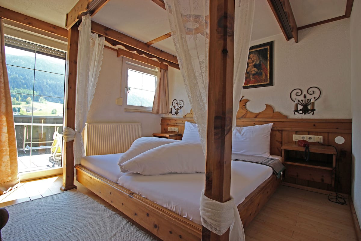 Una cama con dosel y cabecera de madera, una ventana con vistas al exterior, una mesita de noche con una lámpara y una obra de arte colgada en la pared.