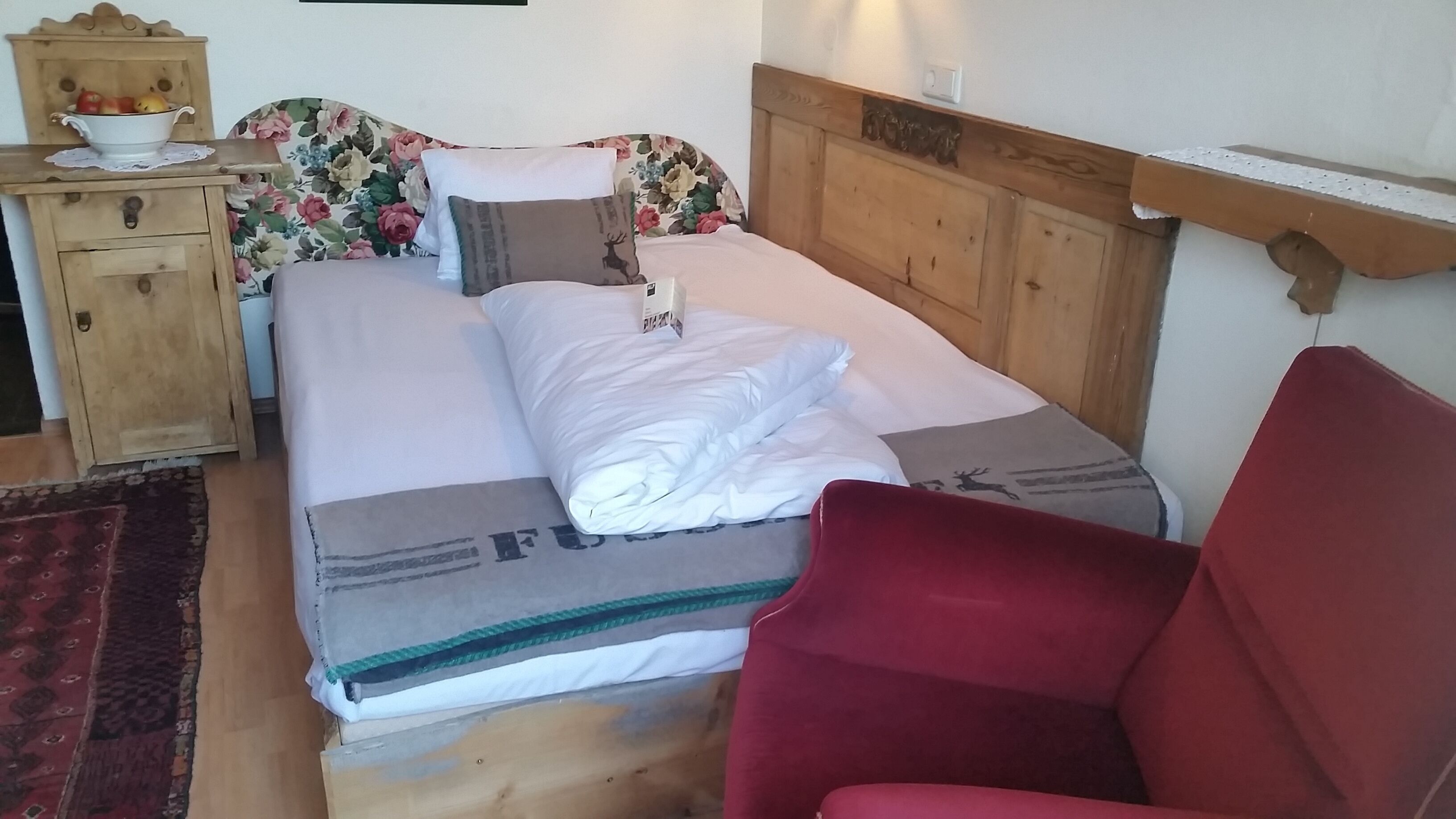 A neatly made bed with a floral headboard, a wooden nightstand with a bowl of fruit, and a red chair.