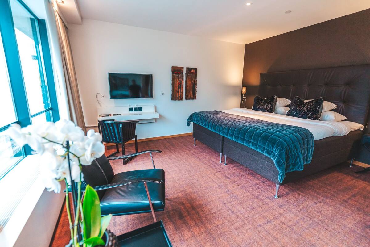 A modern hotel room with a large bed, a desk with a flat-screen TV, a chair, and a potted plant.