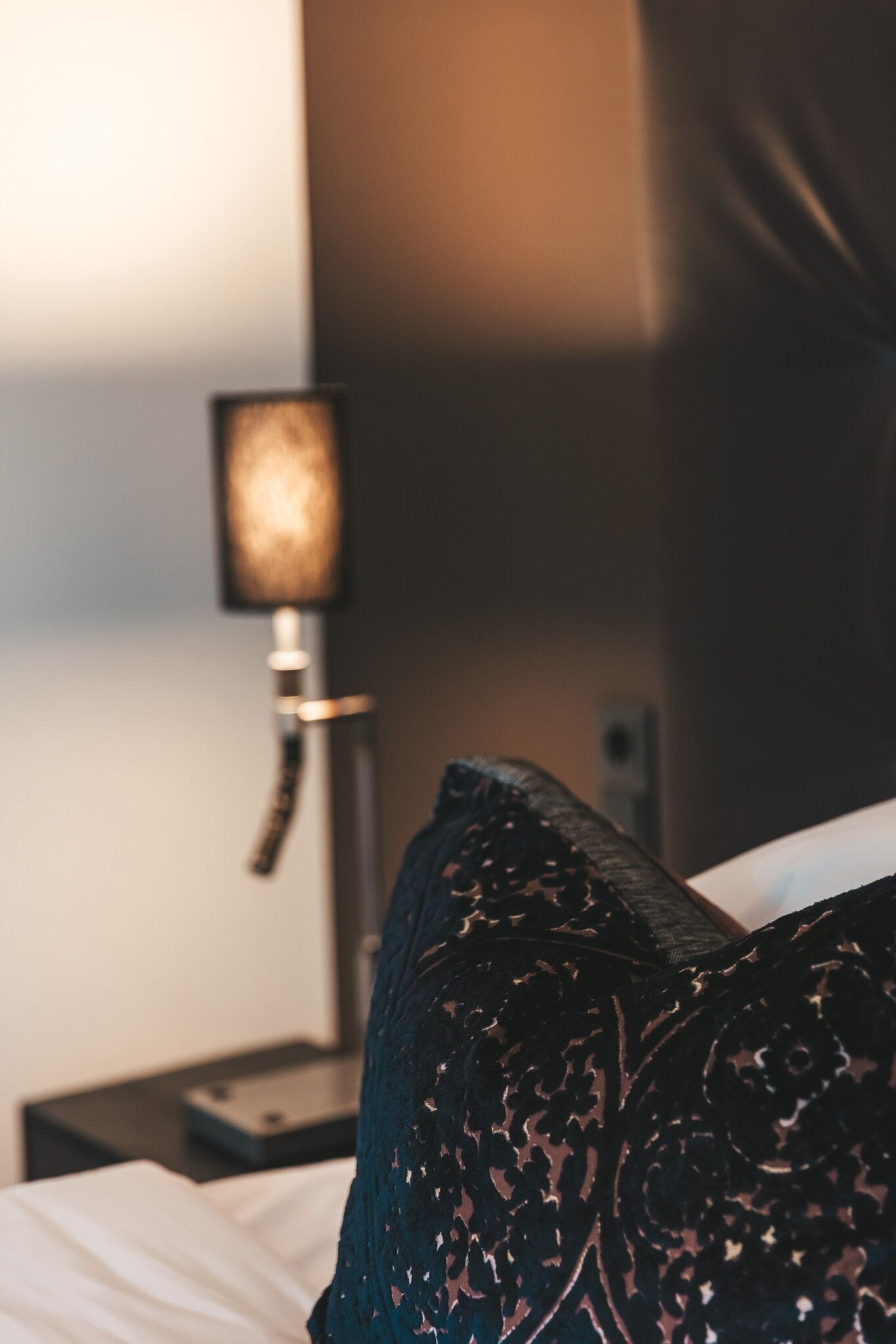 A bed with a dark patterned pillow and a bedside table with a lamp.