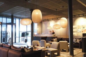 Un hall d’hôtel moderne, doté d’espaces aménagés avec des sièges, de grandes fenêtres et de luminaires suspendus.
