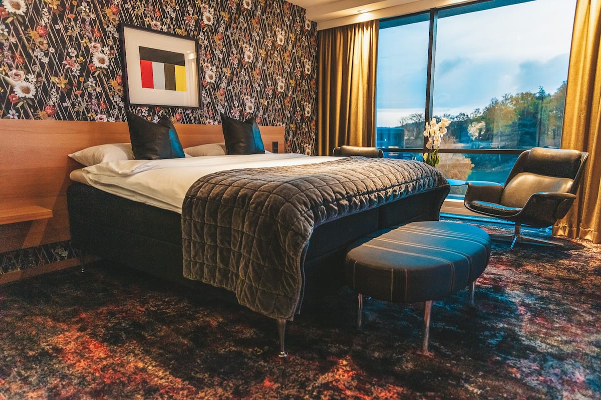 A hotel room with a large bed, a desk, a chair, and a view of the city through large windows.