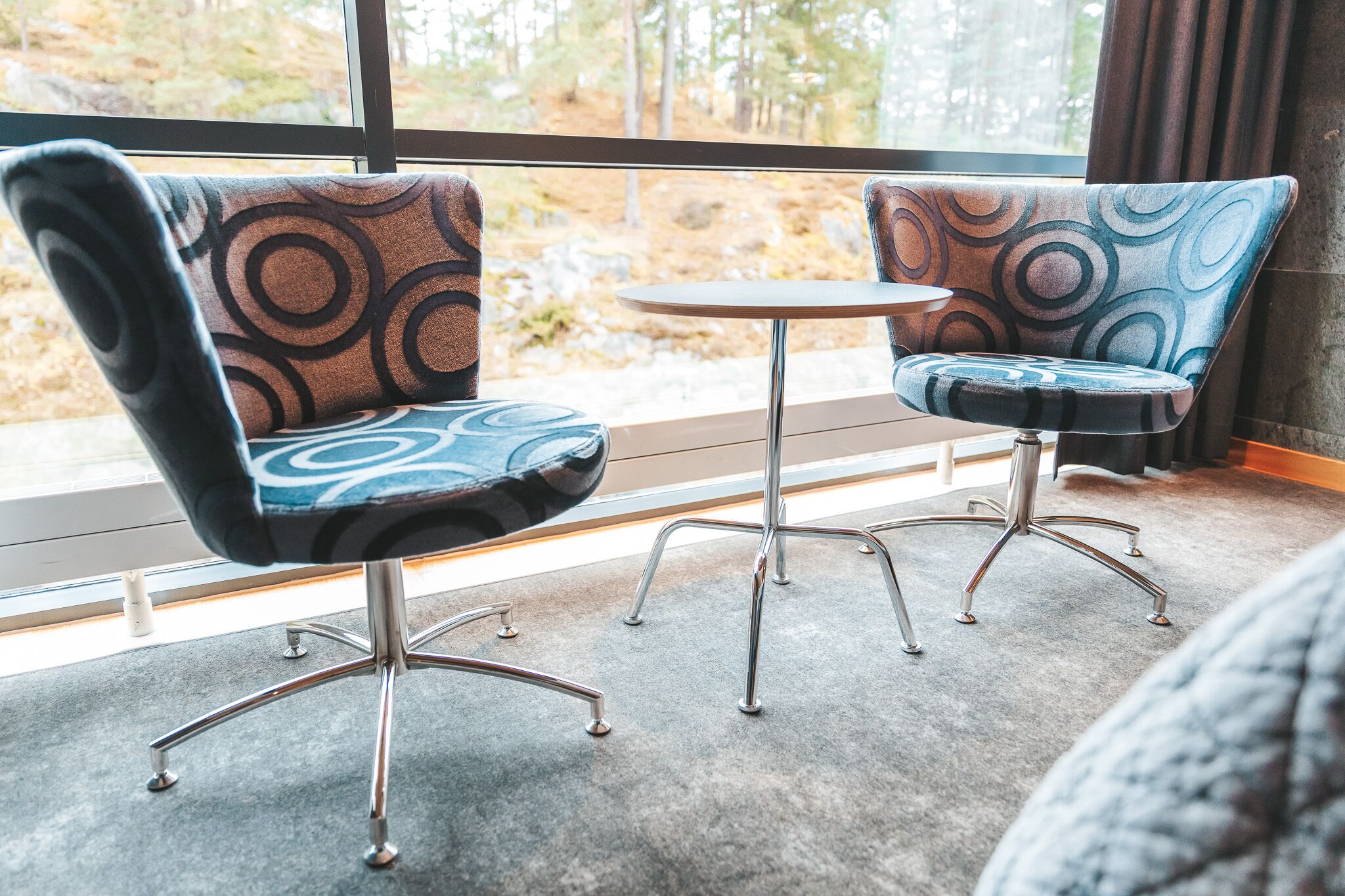 Two modern chairs with a circular pattern upholstery, a small round table, and a view of a forest through large windows.