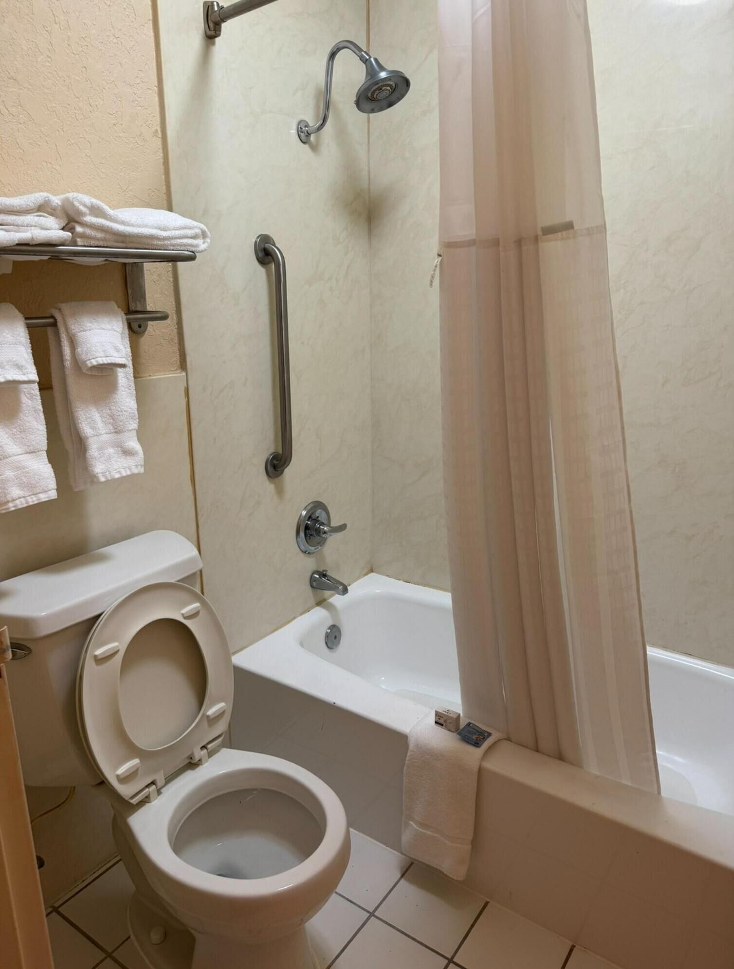 Un baño con inodoro, bañera y ducha. Hay barras de apoyo y un perchero para toallas.