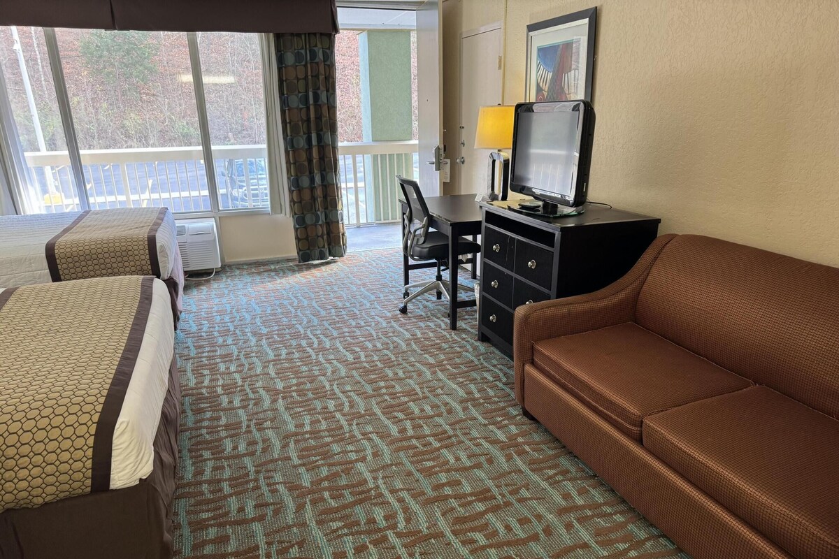 Una habitación de hotel con dos camas, un sofá marrón, un escritorio con una computadora y una ventana con vista a árboles.