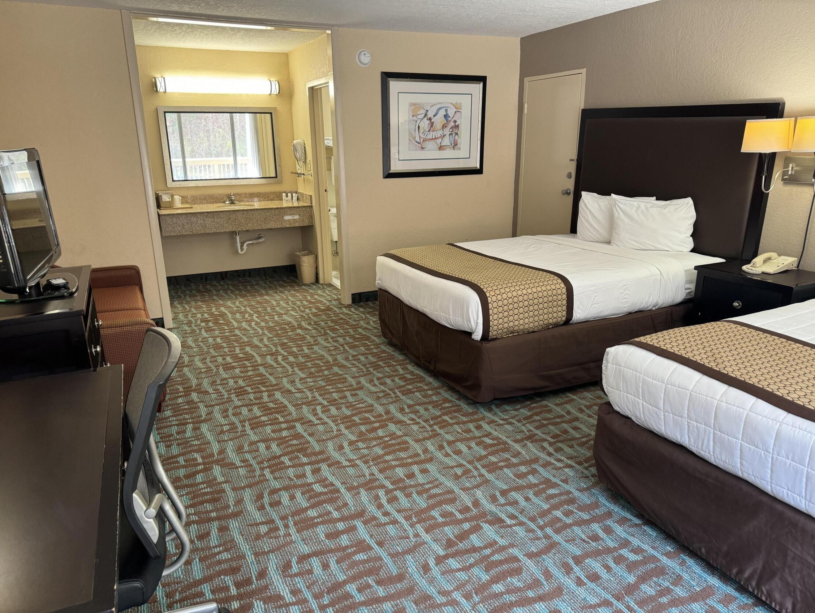 A hotel room with two beds, a desk with a computer, a bathroom with a sink and mirror, and a patterned carpet.
