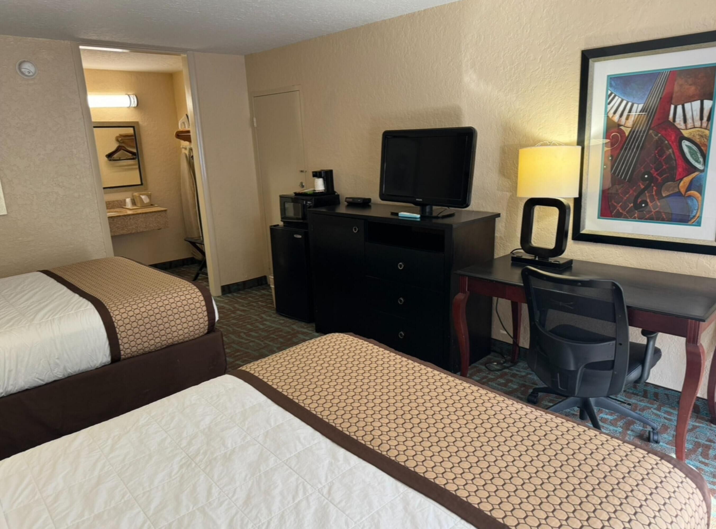 A hotel room with two beds, a desk with a computer and a lamp, a television, and a framed picture on the wall.