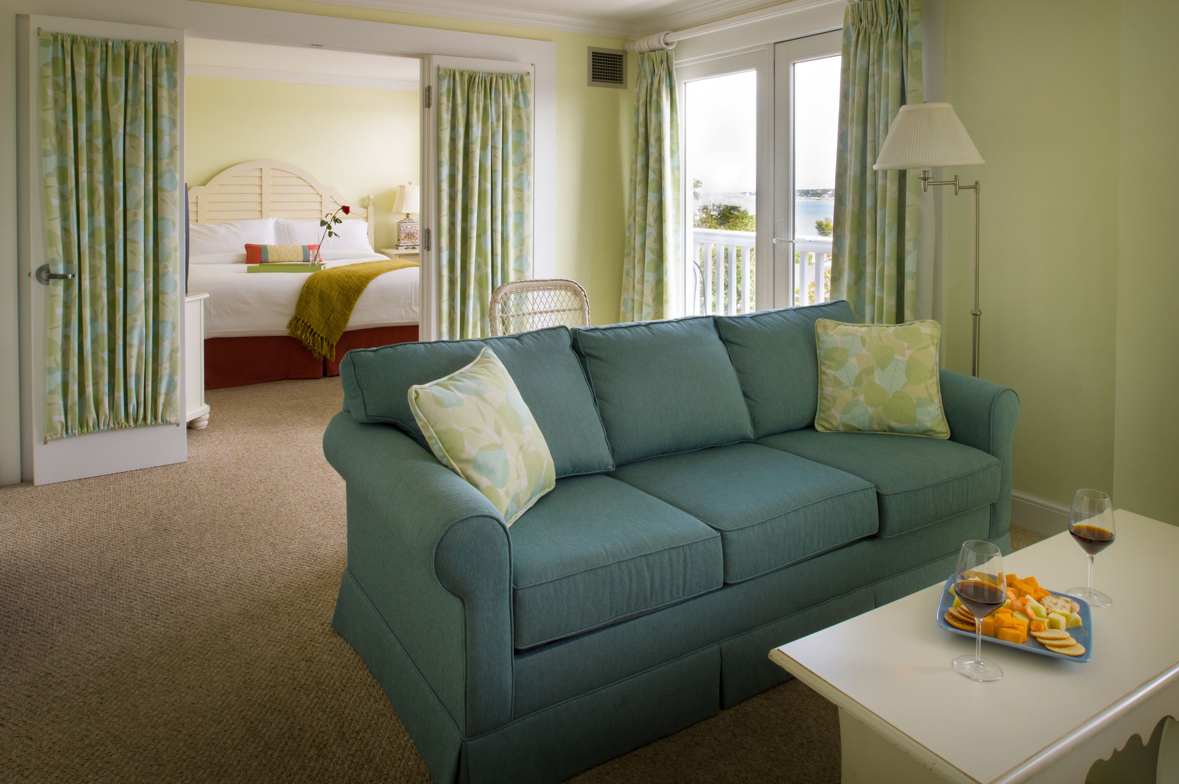 A room with a teal sofa, a bed with a wooden headboard, a dining table with a fruit platter and wine glasses, and a balcony with a view.