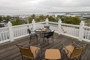 Une terrasse en bois avec une balustrade blanche, une table ronde entourée de chaises, et une vue sur le port de plaisance et la ville en arrière-plan.