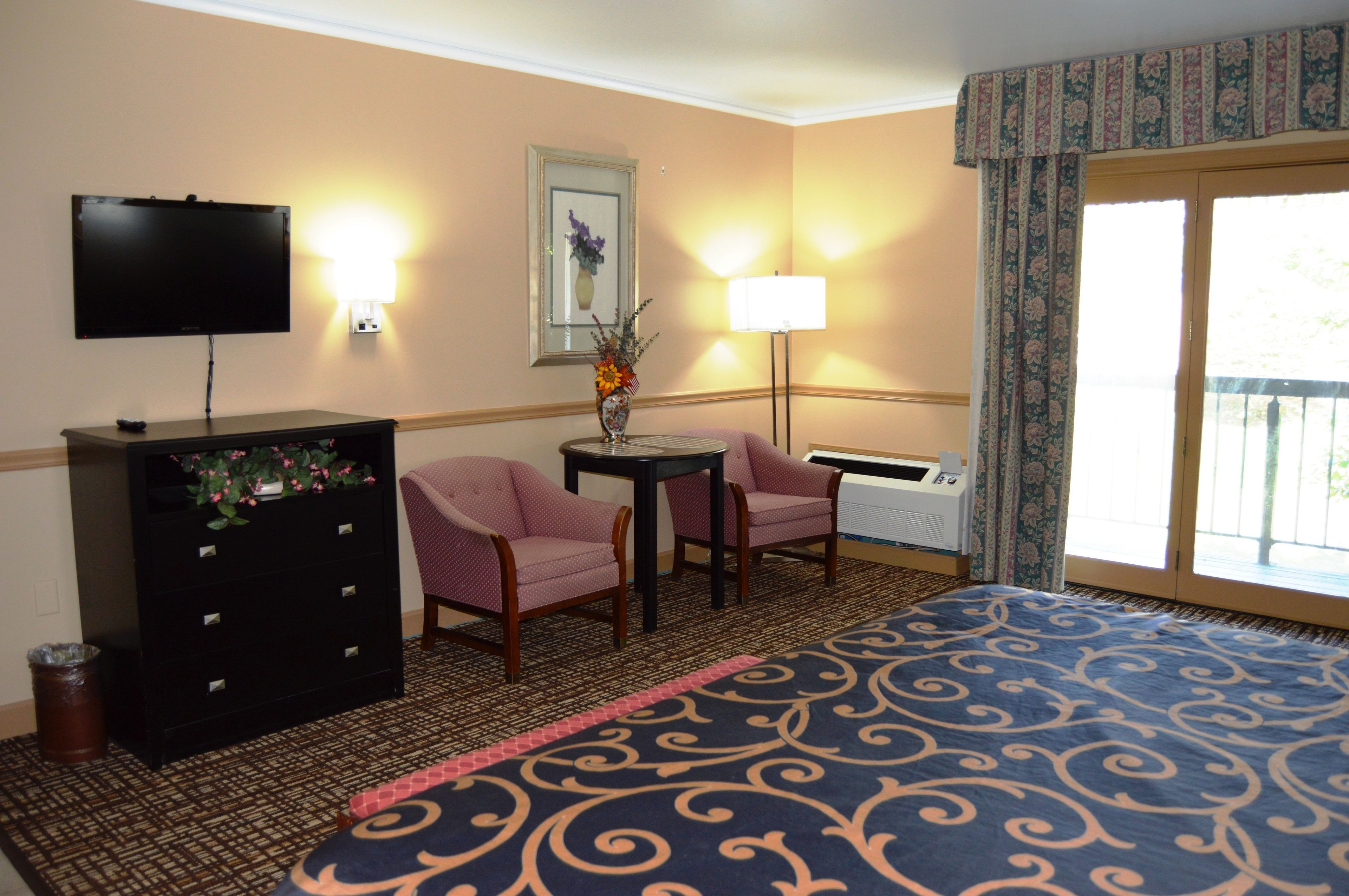 Una habitación de hotel con una cama, dos sillas, un escritorio, un televisor, una lámpara, un cesto de basura y una ventana con cortinas.