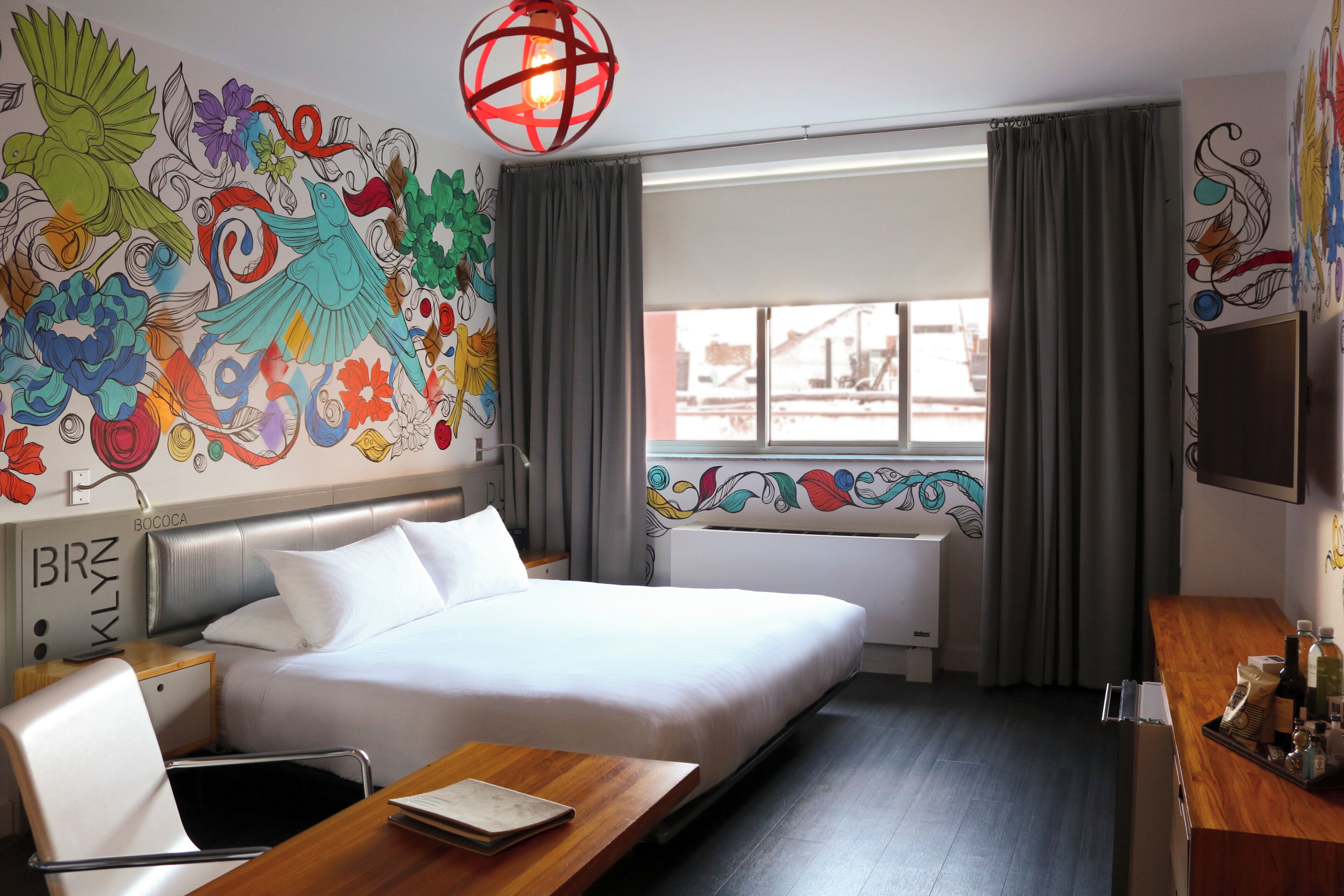 Une chambre d’hôtel comprenant un lit, un bureau, une chaise, une fenêtre avec des rideaux et une fresque colorée sur le mur.