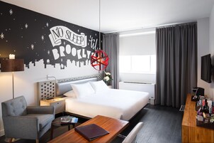 Une chambre d’hôtel comprenant un lit, un bureau, une chaise, une table de chevet, une lampe, un élément de décoration murale et une fenêtre avec des rideaux.