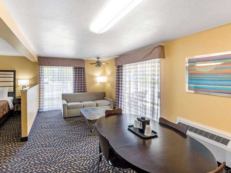 A hotel room with a bed, a sofa, a dining table with chairs, a ceiling fan, and a large window with curtains.