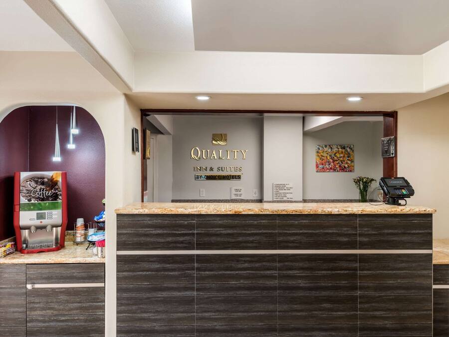 Een hotel receptie met een koffieapparaat, een digitaal scherm en een bordje met de tekst 'QUALITY INN & SUITES BY-CHOICE HOTELS'.