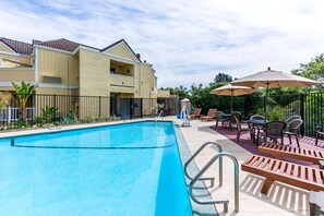 A swimming pool with a metal ladder, adjacent to a building with balconies and a patio area with outdoor seating and umbrellas. - Quality Inn & Suites Capitola By the Sea (Capitola)